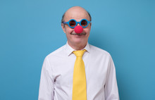 Handsome Senior Man In Clown Nose Yellow Tie And Blue Sun Glasses Ready For Firts Of April, Day Of Fool. Studio Shot On Blue Wall.