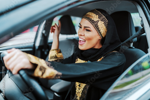 Fotomural Cheerful gorgeous positive muslim woman in traditional wear driving her new car, listening music and singing