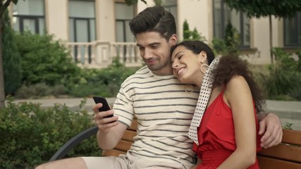 Canvas Print - A smiling couple man and woman are having a video call while sitting on bench at city street