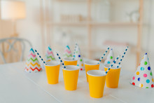 Holiday Hats And Paper Cups With Beverage And Straws On White Table. Festive Event. Birthday Party Celebration Concept. Blurred Background. Nobody On Picture