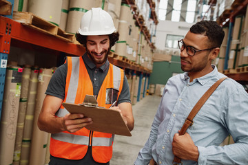 Storehouse factory worker discussing document with list of goods with warehouse manager holding clipboard writing using pen