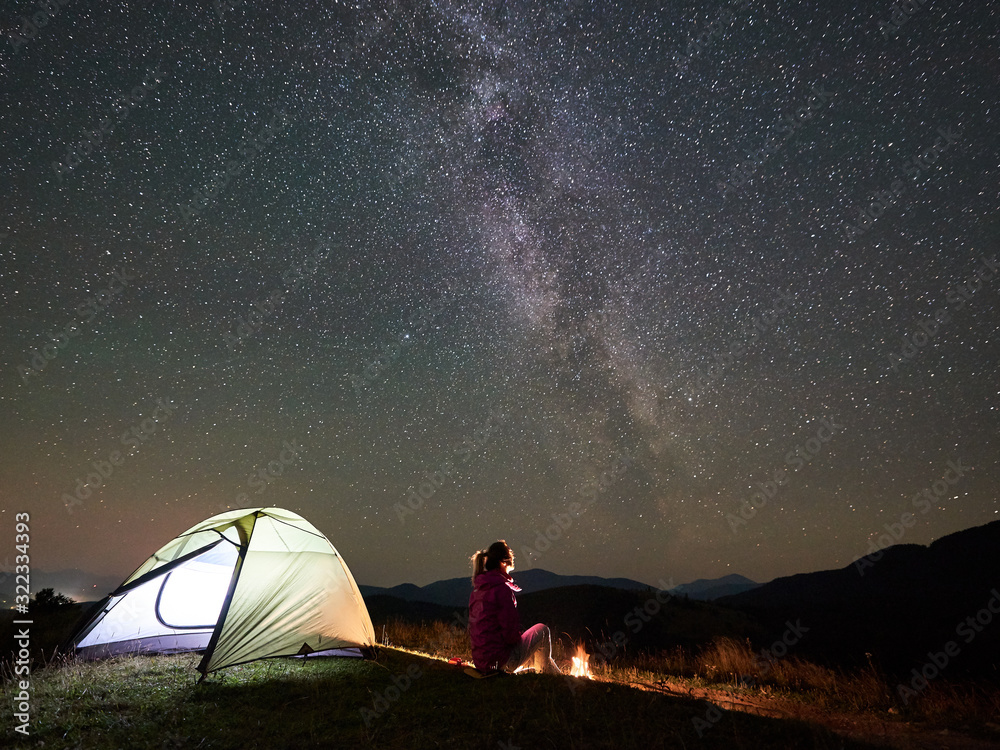 Female camper having a rest at summer night camping in the mountains beside bonfire, glowing tourist tent. Young woman enjoying view of night sky full of stars and Milky way. Tourism adventure concept - obrazy, fototapety, plakaty 