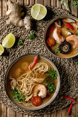 Variety of asian soups. Traditional spicy Thai tom yum kung and noodles soup with shiitake mushrooms, prawns, ingredients above on straw wicker napkin over wooden plank background. Flat lay, space.