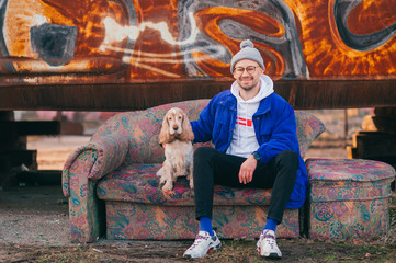 Stylish young man in blue old fashioned jacket, white hoodie and modern sneakers siiting on old couch with best friend pet - cocker spaniel dog over rusty metal graffiti wall on background.