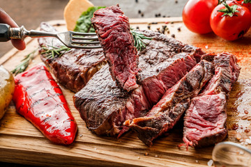 Wall Mural - Hands cut grilled tomahawk meat medium rare or rib eye steak on wooden cutting board with grilled vegetables on dark background, close up