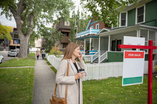 Woman With Coffee Talking On Smart Phone And Looking At House For Sale In Neighborhood