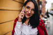 © Look! - Close-up portrait of smiling brunette girl with red lips talking on phone. Outdoor photo of winsome female secretary in glasses posing on the street with smartphone.