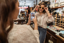 Young Woman Trying On Sunglasses With Friend Taking Photo On Smart Phone
