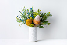 A Elegant Floral Arrangement In A White Vase On A White Table With A White Background. Flower Bunch Includes Blue And Yellow Flowers, Pink And Yellow Australian Native Banksias And Eucalyptus Leaves.
