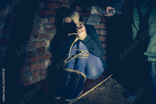 Fotografía irl with her hands and feet tied is sitting in the basement and screaming, and a man in rubber boots is standing next to her with an axe in his hands