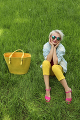 A young woman with a bag is on the background of green grass in yellow pants and a pink jacket with sandals.Urban summer style.