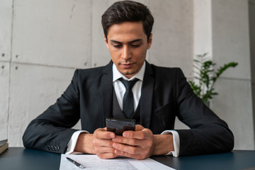 Serious attractive young businessman in dark suit holding smartphone while sitting at loft office table. Concept of internet and technology