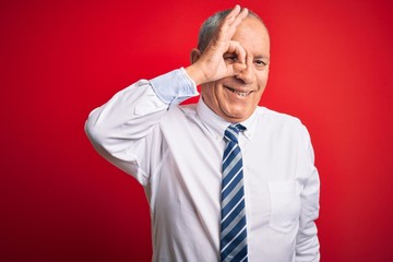 Wall Mural - Senior handsome businessman wearing elegant tie standing over isolated red background doing ok gesture with hand smiling, eye looking through fingers with happy face.