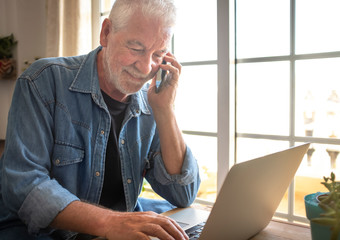Close up of a senior adult man  with beard and white hair working at home using the laptop. Talking at the cellphone. Bright light from window.  One caucasian people
