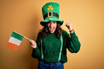 Beautiful patriotic curly hair woman wearing hat holding irish flag celebrating saint patricks day screaming proud and celebrating victory and success very excited, cheering emotion