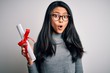 Young beautiful chinese woman holding diploma standing over isolated white background scared in shock with a surprise face, afraid and excited with fear expression