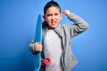 Poster - Young little boy kid skateboarder holding modern skateboard over blue isolated background annoyed and frustrated shouting with anger, crazy and yelling with raised hand, anger concept