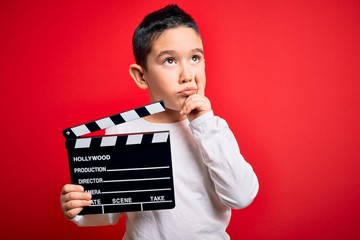 Wall Mural - Young little boy kid filming video holding cinema director clapboard over isolated red background serious face thinking about question, very confused idea