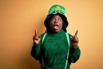 Wall Mural - Plus size african american woman with braids wearing green hat with clover on st patricks day amazed and surprised looking up and pointing with fingers and raised arms.