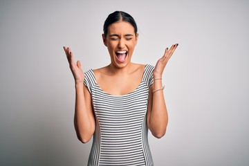 Young beautiful brunette woman wearing casual striped dress over isolated white background celebrating mad and crazy for success with arms raised and closed eyes screaming excited. Winner concept