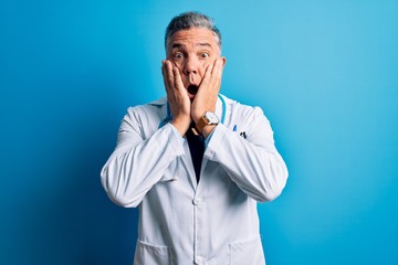 Poster - Middle age handsome grey-haired doctor man wearing coat and blue stethoscope afraid and shocked, surprise and amazed expression with hands on face