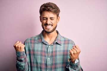 Young handsome man with beard wearing casual shirt standing over pink background excited for success with arms raised and eyes closed celebrating victory smiling. Winner concept.