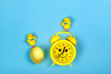 Retro Yellow Alarm Clock With Drawn Smiling Face And Toys Chicken And Colored Egg On Blue Background. Easter Concept.