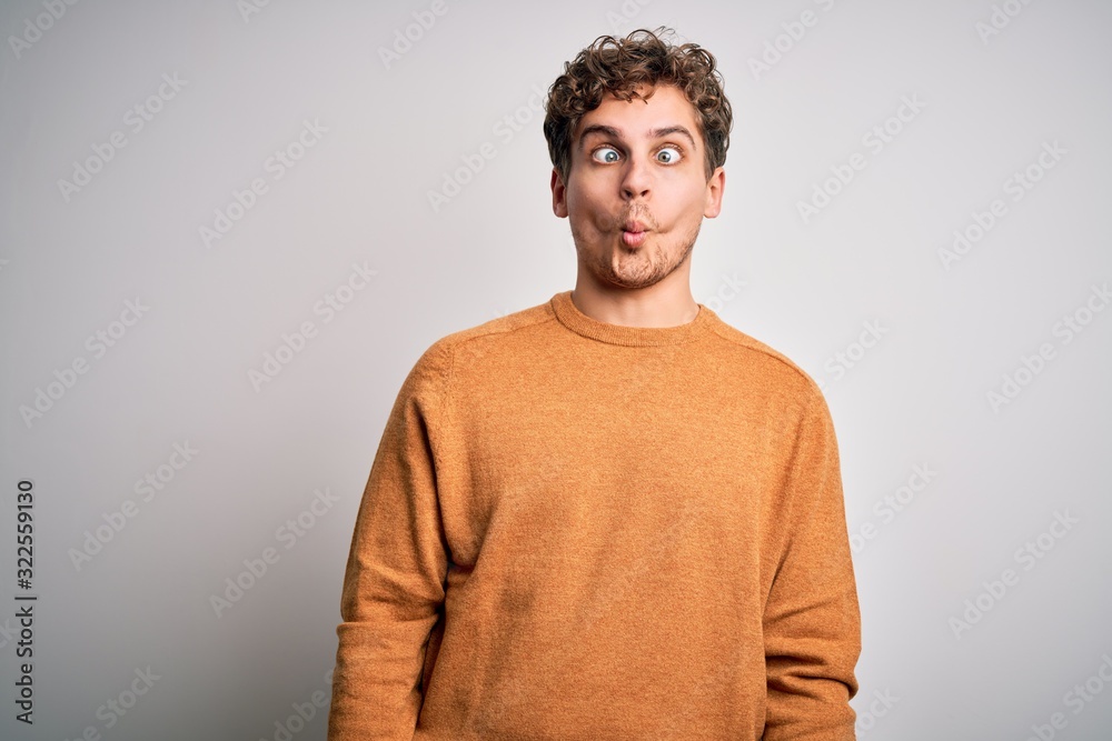 Young blond handsome man with curly hair wearing casual sweater over white background making fish face with lips, crazy and comical gesture. Funny expression.
