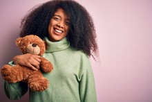 Young African American Woman With Afro Hair Hugging Teddy Bear Over Pink Background With A Happy Face Standing And Smiling With A Confident Smile Showing Teeth