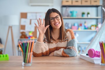 Young beautiful teacher woman wearing sweater and glasses sitting on desk at kindergarten showing and pointing up with fingers number five while smiling confident and happy.