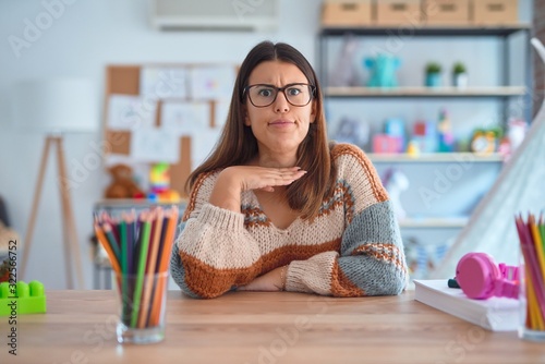 Fotomural Young beautiful teacher woman wearing sweater and glasses sitting on desk at kin
