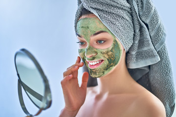 Smiling young happy healthy female with green face clay mask and small round mirror during spa day at home. Face skin care