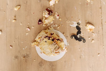 Smashed First Birthday Cake On Floor Flat Lay Photo. Destroyed Delicious Creamy Celebrate Biscuit Dessert Heaped Mess And Paper Moustache Decoration. Remains Pieces Horizontal Photography