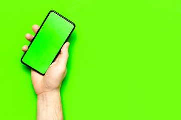 Male hands hold a modern black smartphone with green blank screen on neon green background flat lay top view. Modern technology, phone, gadget in hands, touch screen, template for your design. Mockup