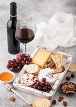 Glass And Bottle Of Red Wine With Selection Of Various Cheese In Wooden Box And Grapes On Light Table Background. Blue Stilton, Red Leicester And Brie Cheese With Cheddar And Nuts With Honey.