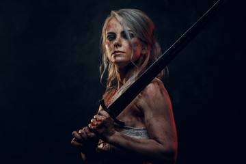 Wall Mural - Naked fantasy woman warrior wearing rag cloth stained with blood and mud posing with a sword. Studio photo on a dark background. Cosplayer as Ciri from The Witcher