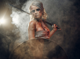 Wall Mural - Woman warrior wearing rag cloth stained with blood and mud posing with a sword and shield. Studio photo on a dark background with smoke. Cosplayer as Ciri from The Witcher