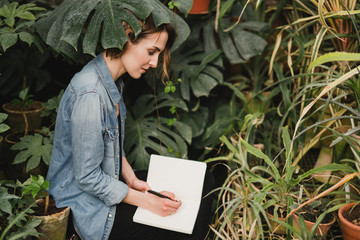 Young beautiful caucasian woman in glass greenhouse among colorful greenery leaves and flowers. Art portrait of a girl with notebook taking notes. Gardener, writer, blogger, nature lover, inspiration.