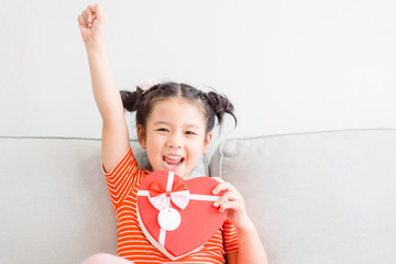 Wall Mural - Little asian girl raised hands smile and excited and holding red gift box heart shape in sofa at home on white wall background.Child girl holding gift box in Valentine, Christmas and New year concept.