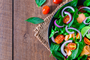 Salmon salad, view from above with fresh tomato, onion, spinach, lettuce and basil in black bowl on wooden background. Home made concept. Diet, vegetable for tasty and healthy. Top view, copy space.