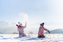 Cheerful Little Girls In Outwear Having Fun And Throwing Snow In Sunlight Standing Outdoors