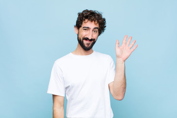 Wall Mural - young crazy bearded man smiling happily and cheerfully, waving hand, welcoming and greeting you, or saying goodbye against flat wall