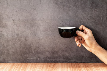 A man's hand holding a black coffee mugs on wooden table with old gray concrete walls.