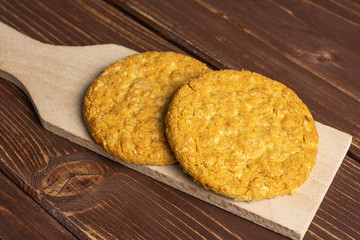 Group of two whole sweet golden oat cookie on small wooden cutting board on brown wood