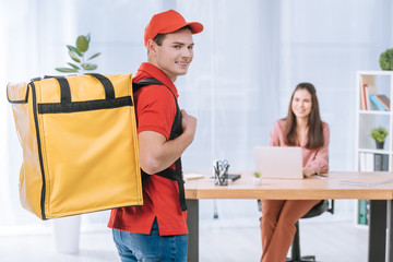 Wall Mural - Selective focus of delivery man with thermo backpack smiling at camera with businesswoman at table in office