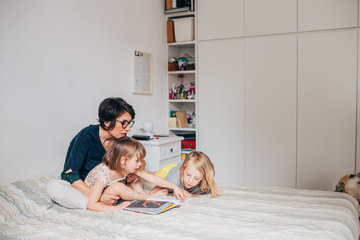 Mother of two female children sitting on bed reading book together