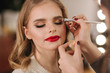 © Aleksandr - Makeup artist applies eye shadow, perfect evening makeup. Beauty blond hair girl with perfect skin and red lipstick