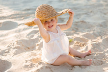 Baby Girl In White Clothes And A Straw Hat Sits On The White Sand On The Beach In Summer