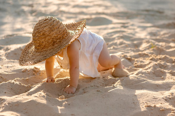 baby girl in white clothes and a straw hat sits on the white sand on the beach in summer