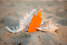 Two Large Seashells, Sunscreen In An Orange Tube And A Straw Hat Lie On A White Sandy Beach